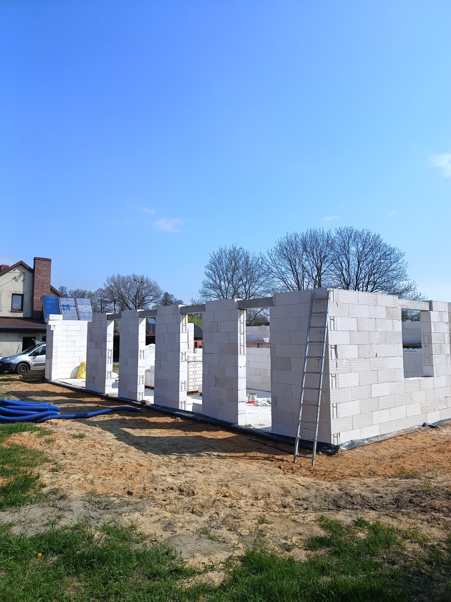 Wznoszone ściany parterowego budynku z białych bloczków silikatowych, widoczna drabina oparta o ścianę, niebieskie rury na ziemi, w tle dom jednorodzinny i błękitne niebo.