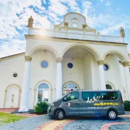 Szary van z numerem telefonu na tle eleganckiego budynku z kolumnami i rzeźbami, w tle para ubrana na galowo.