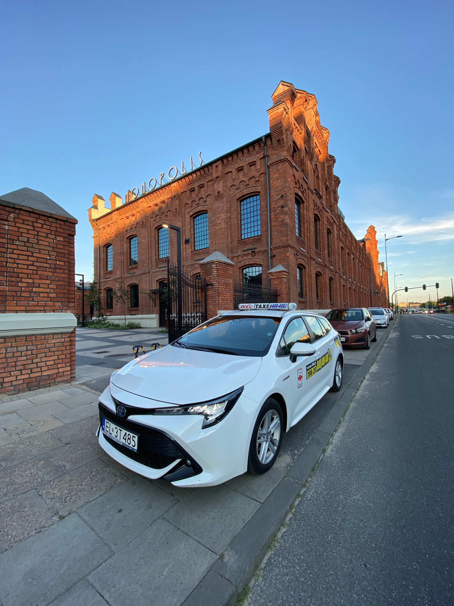 Biała taksówka Toyota Corolla zaparkowana przed ceglanym budynkiem Monopolis w Łodzi, widoczny numer rejestracyjny EL 3T485.
