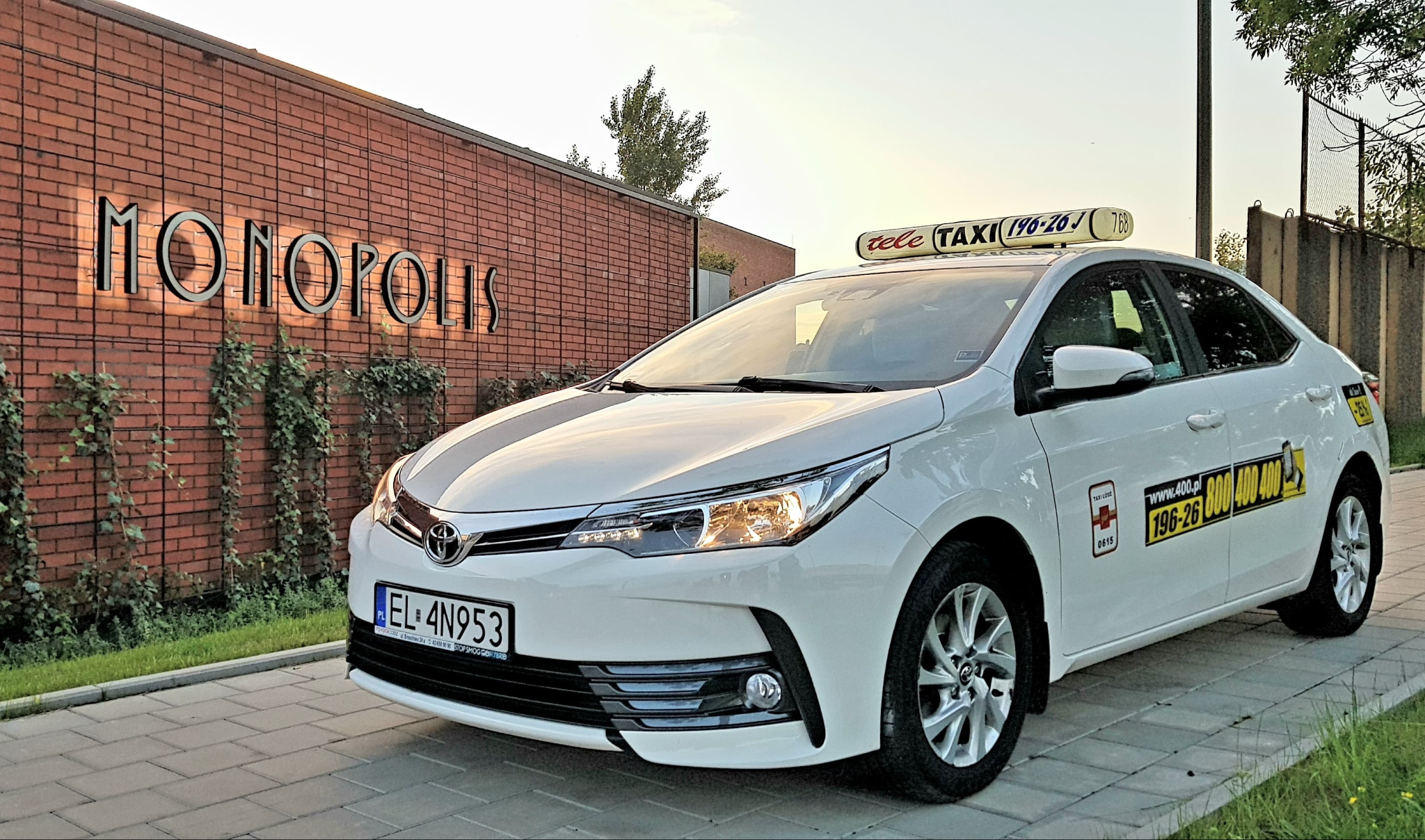 Biała taksówka Toyota Corolla z numerem rejestracyjnym EL 4N953 na tle ceglanej ściany z napisem MONOPOLIS, z widocznymi numerami telefonu i logo Taxi Łódź.