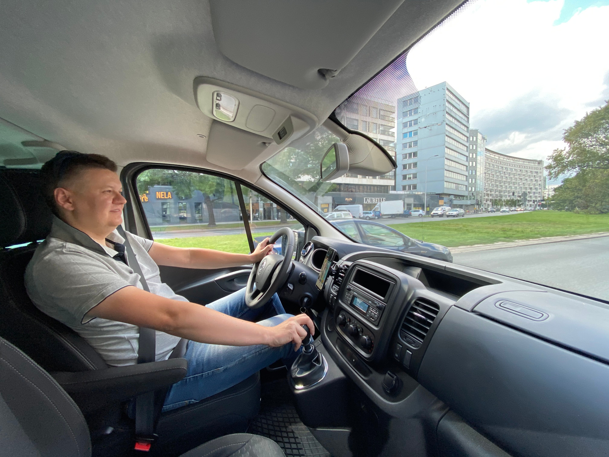 Kierowca w trakcie jazdy busem Opel Vivaro, widok z wnętrza kabiny na ulicę z budynkami w tle.