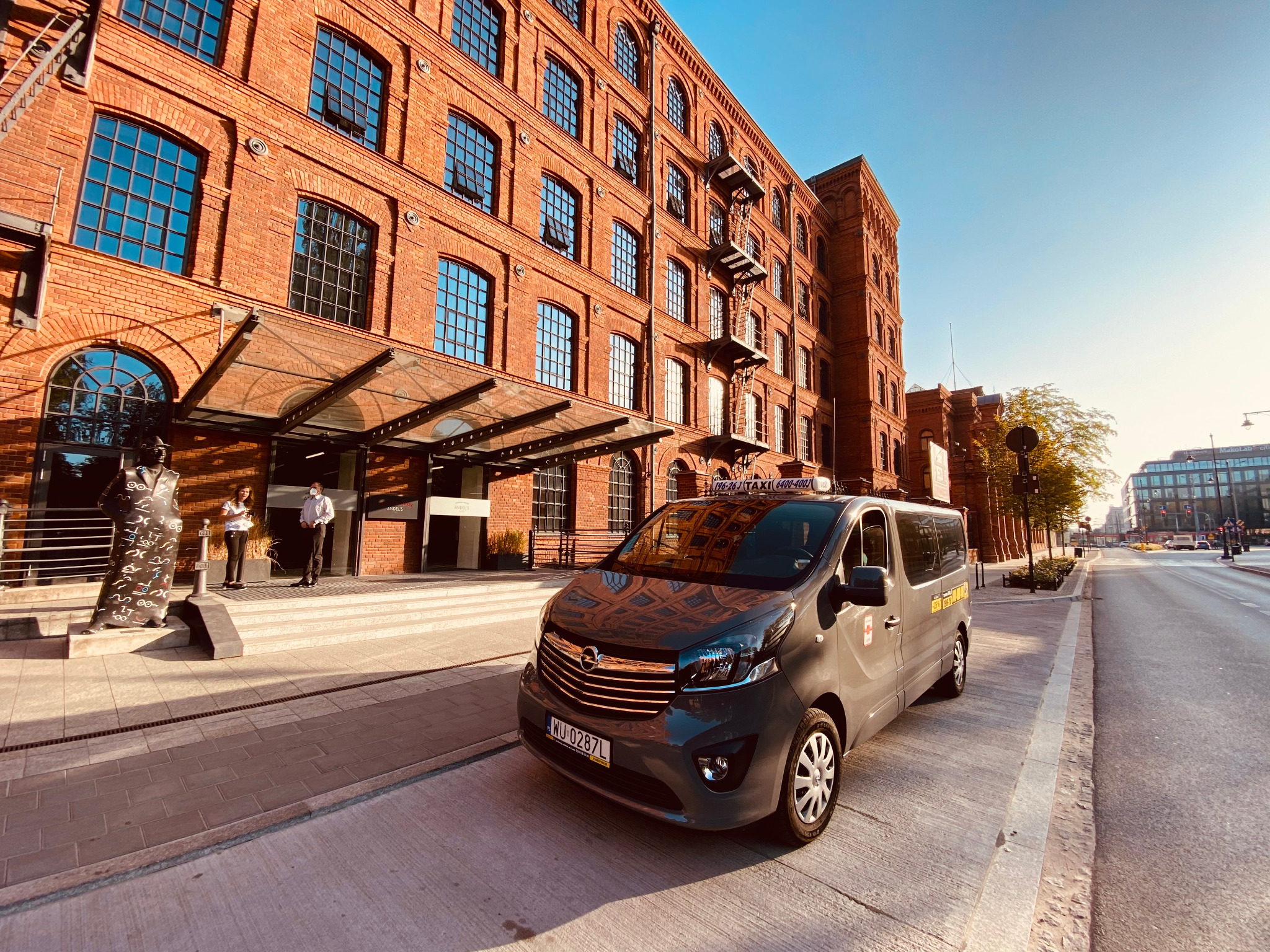 Szary van taxi z numerem rejestracyjnym WU 0287L zaparkowany przed budynkiem z czerwonej cegły z przeszklonym wejściem i rzeźbą.
