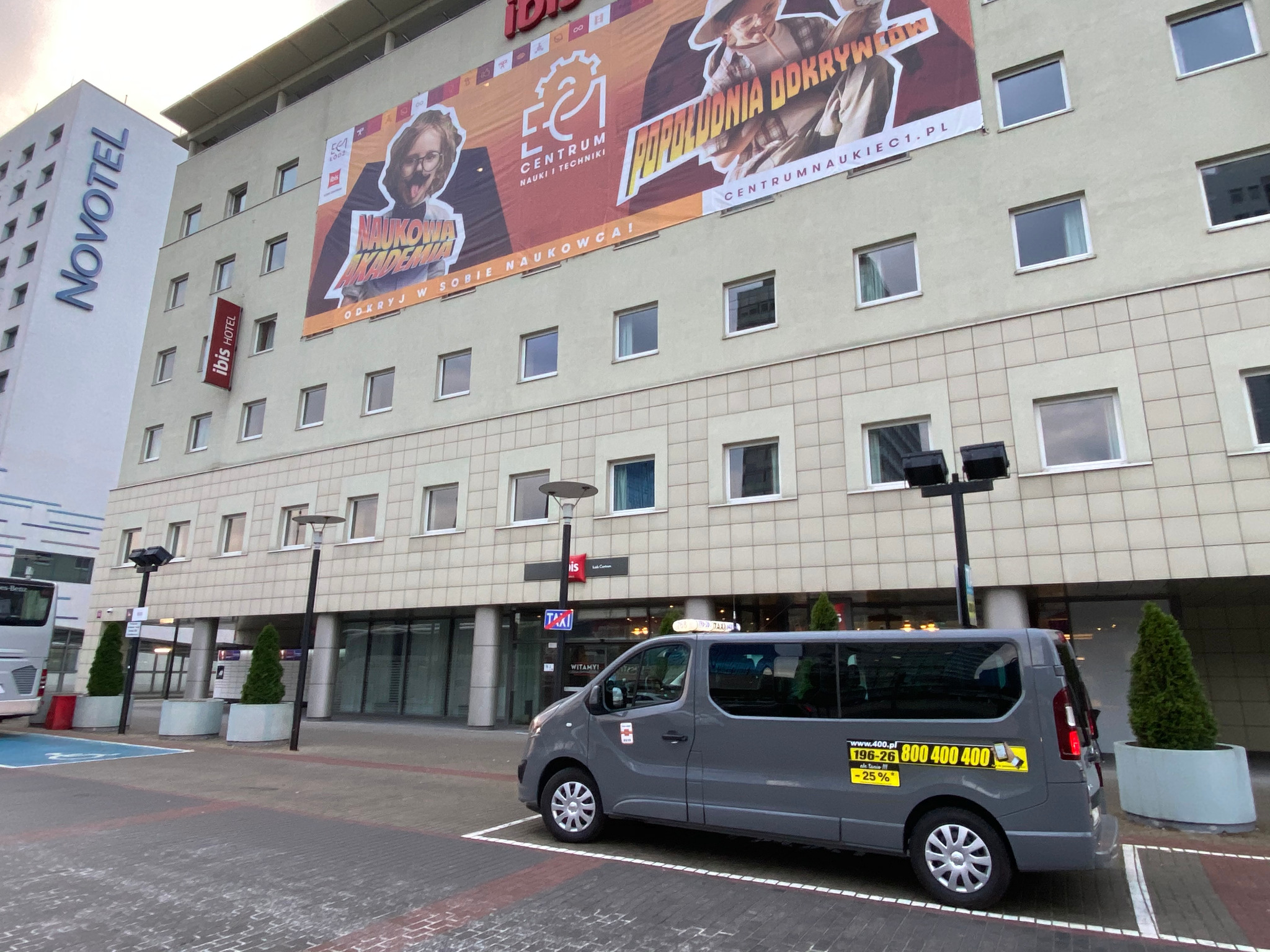 Szara taksówka typu van zaparkowana przed budynkiem Ibis Hotel i Novotel z reklamą Centrum Nauki i Techniki, widoczny numer telefonu i napis 'ale tania !!! -25%'.