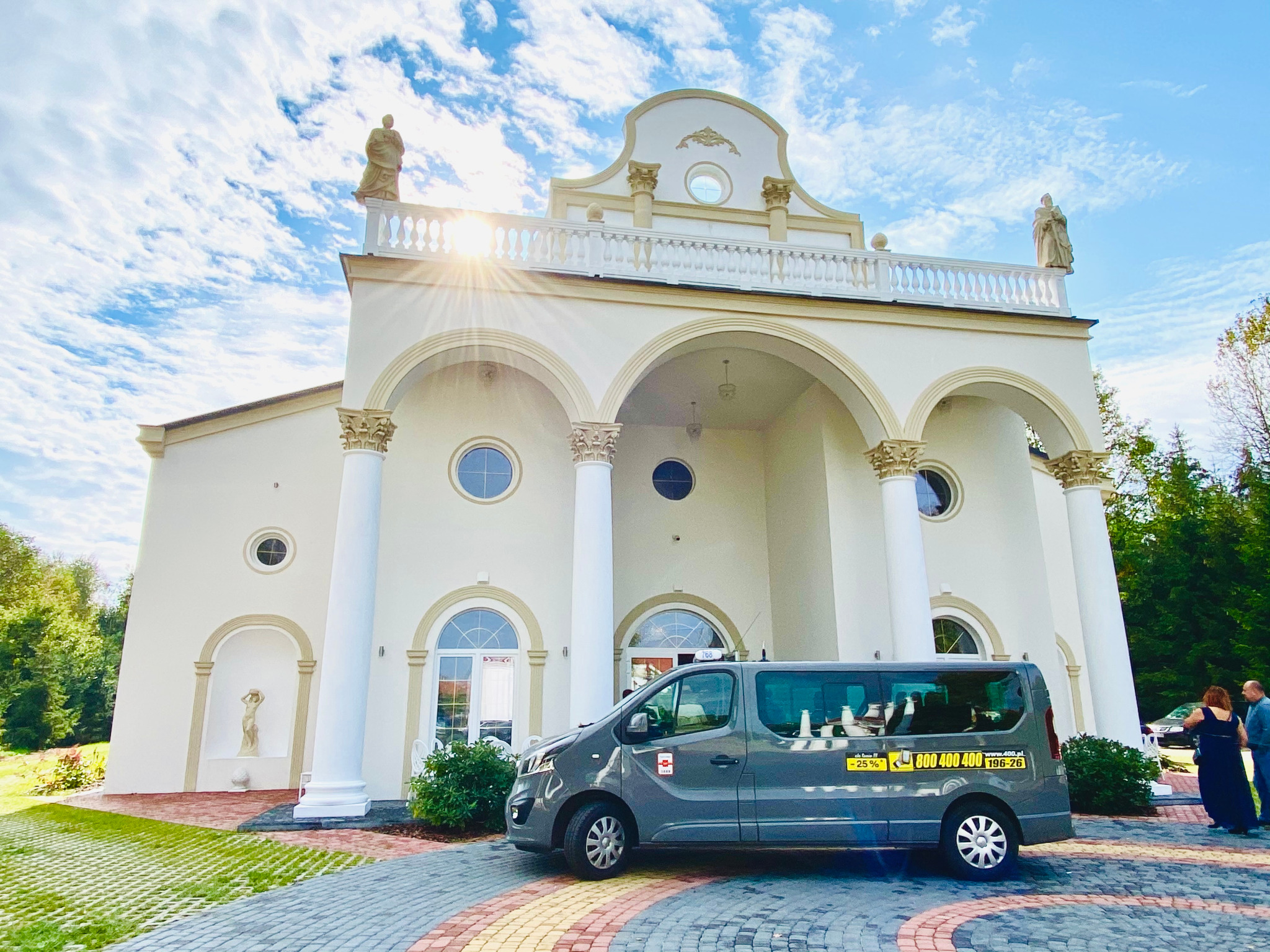 Szary van z numerem telefonu na tle eleganckiego budynku z kolumnami i rzeźbami, w tle para ubrana na galowo.