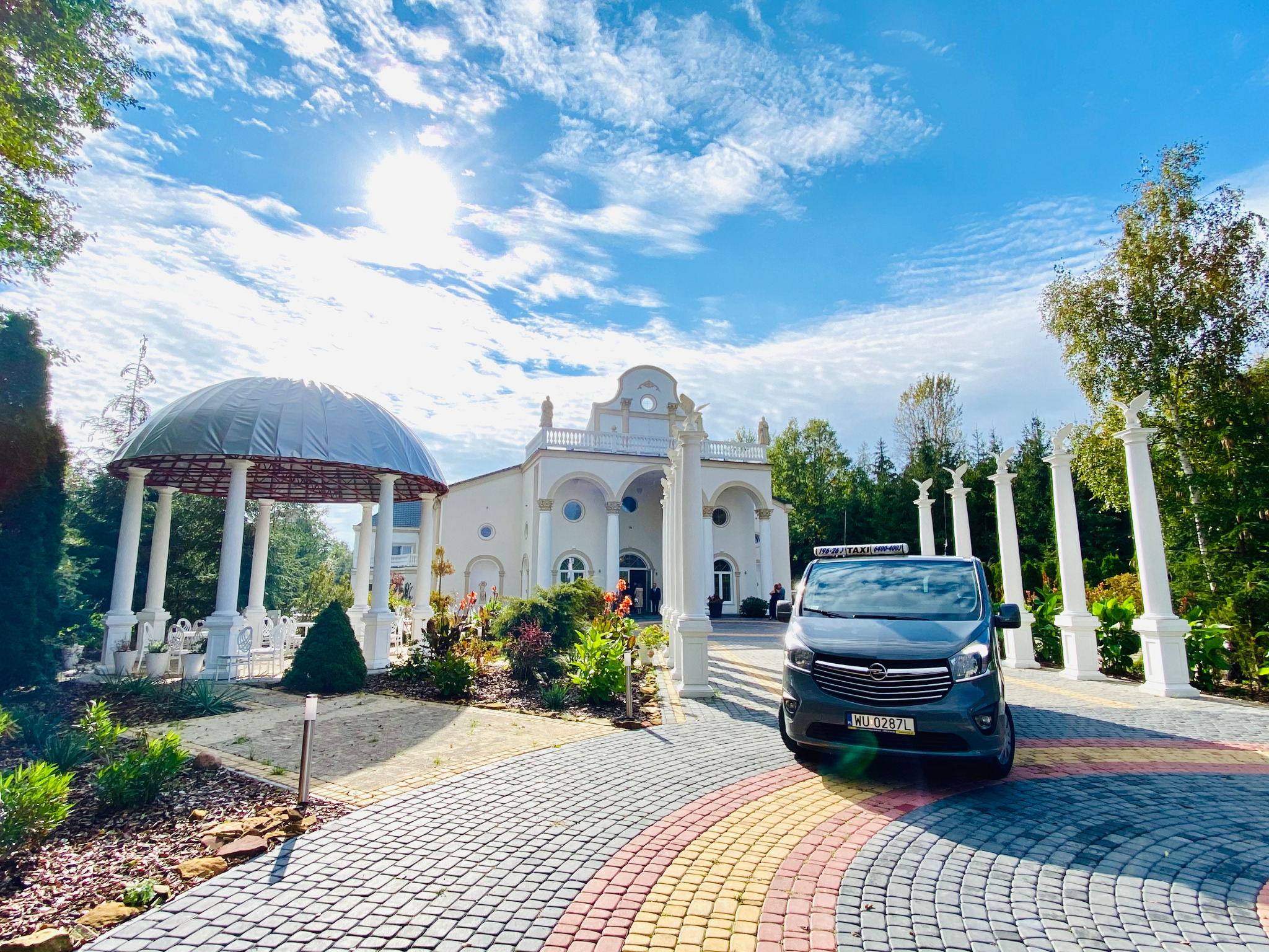 Ciemnoszary van z napisem TAXI na dachu, zaparkowany na kolorowej kostce brukowej przed reprezentacyjnym, białym budynkiem z kolumnami i kopułą w słoneczny dzień.