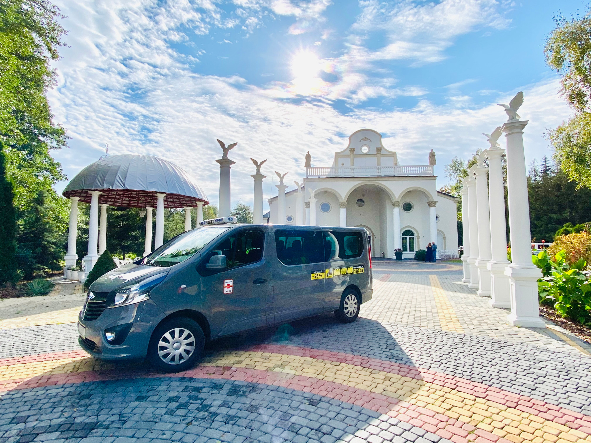 Szary van z oznaczeniami firmy przewozowej zaparkowany na kolorowej kostce brukowej przed białym budynkiem z kolumnami i rzeźbami orłów, w tle altana i błękitne niebo.