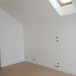 Wnętrze poddasza po remoncie: świeżo pomalowane białe ściany, jasna podłoga, okno dachowe i widoczne punkty instalacji elektrycznej.