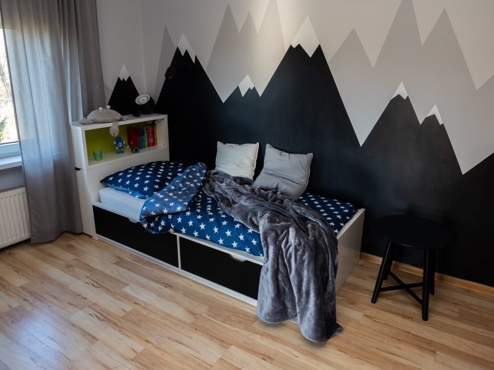 Pokój dziecięcy z łóżkiem z szufladami, pościelą w gwiazdki i ścianą pomalowaną w motyw górski w odcieniach szarości i czerni. Obok łóżka stoi mały, czarny stolik.