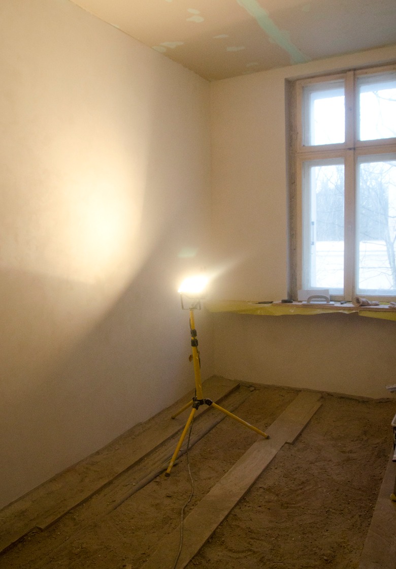 Pokój w trakcie prac wykończeniowych, ściany przygotowane pod malowanie, podłoga z piaskiem i resztkami materiałów, żółty reflektor budowlany oświetla wnętrze, okno zabezpieczone folią malarską.