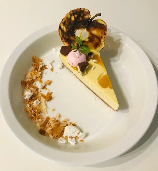 Kawałek sernika z brzoskwiniami, dekorowany różową bezą, czekoladą, miętą i karmelizowanym jabłkiem na białym talerzu z posypką z ciasta francuskiego i bezy.