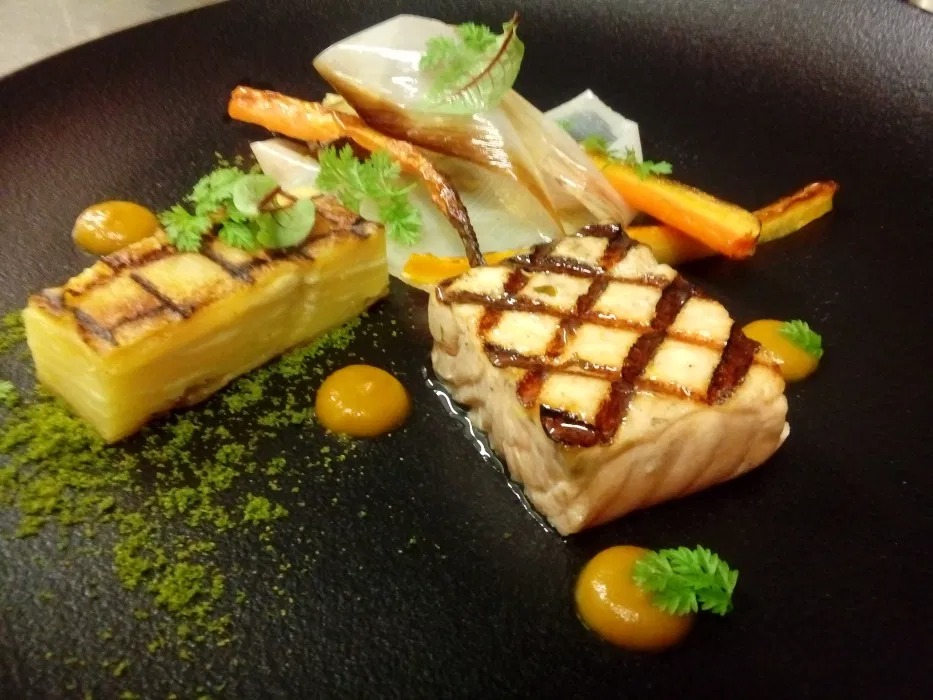 Elegancko podane danie z grillowaną rybą, zapiekanką ziemniaczaną i karmelizowanymi warzywami na czarnym talerzu, udekorowane zielonym pudrem i sosem.