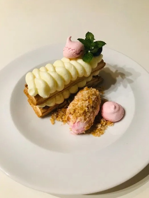 Elegancko podany deser: warstwowy kremowy tort z bezą, posypką i miętą na białym talerzu.