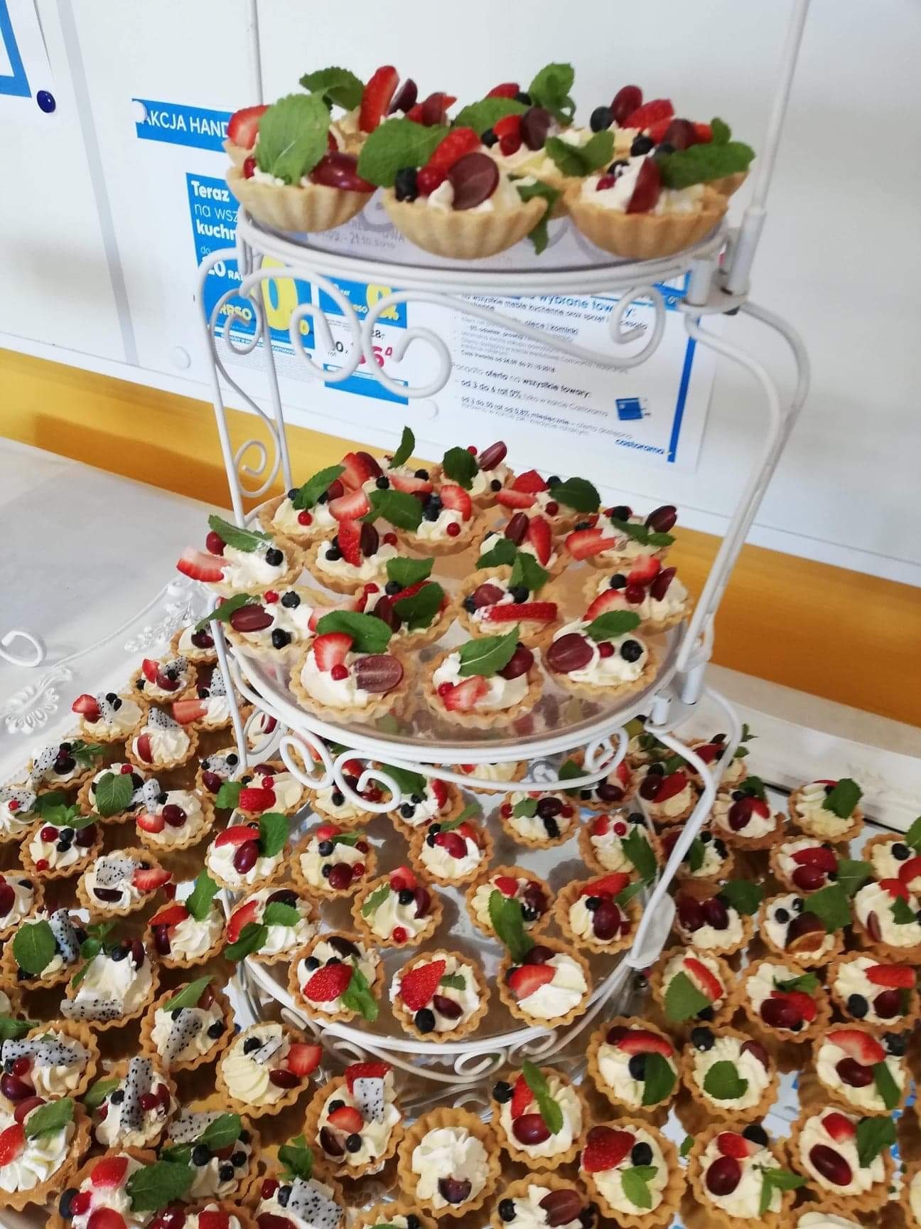 Elegancka, trzypoziomowa patera z mini tartaletkami wypełnionymi kremem i dekorowanymi świeżymi truskawkami, winogronami, borówkami i listkami mięty, ustawiona na srebrnej tacy.