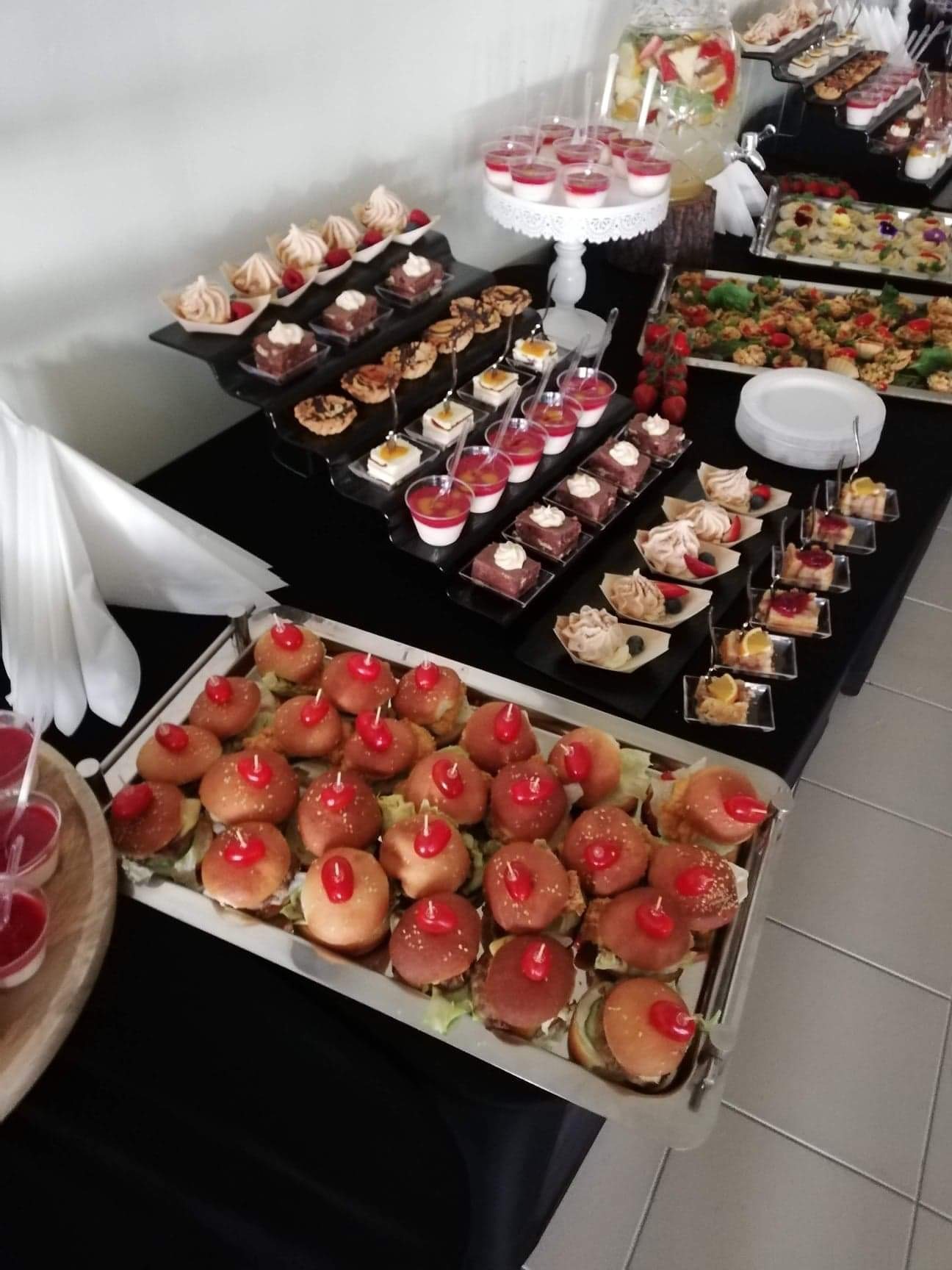 Stół cateringowy z mini burgerami z czerwonymi dekoracjami, deserami w szklankach i na tackach, oraz napojem owocowym w dystrybutorze, przygotowany na czarnym obrusie.