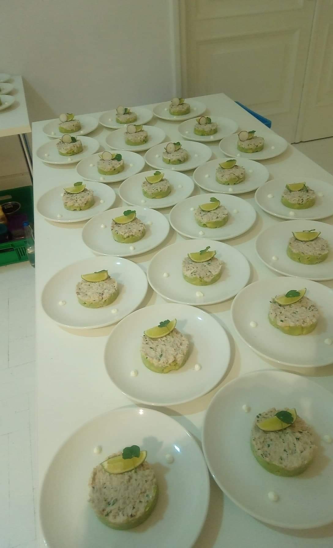 Rzędy białych talerzy z przystawkami: spód z awokado, pasta rybna, plasterek limonki i listek mięty, ustawione na białym stole.