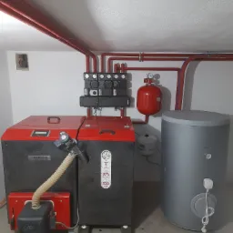 Kotłownia z piecem na paliwo stałe, zbiornikiem akumulacyjnym i rozbudowaną instalacją hydrauliczną z czerwonymi rurami. Piec posiada oznaczenia 'Genesis' i 'ECO design', a na ścianie widoczny jest...