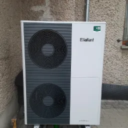 Biała pompa ciepła Vaillant aroTHERM plus zamontowana na betonowych bloczkach z widocznym żółtym wiertłem udarowym, przy ścianie budynku o szorstkiej fakturze.