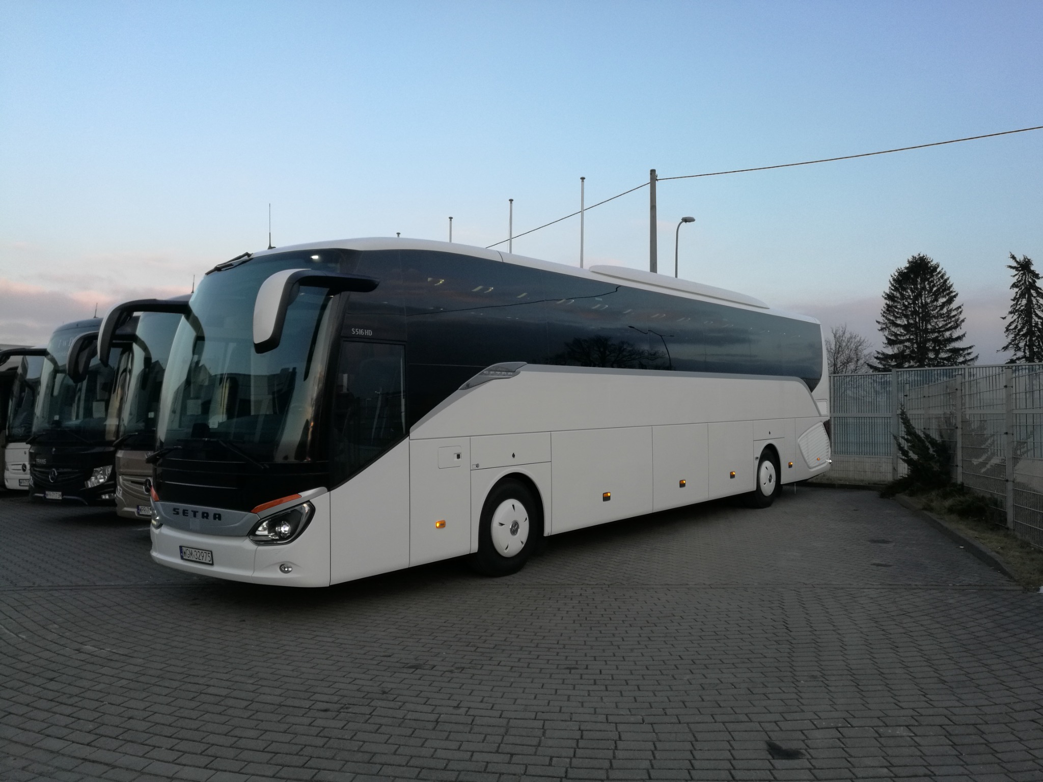 Nowoczesny, biały autokar Setra S516HD zaparkowany na brukowanym placu, z rzędem innych autokarów w tle, sfotografowany o zmierzchu.
