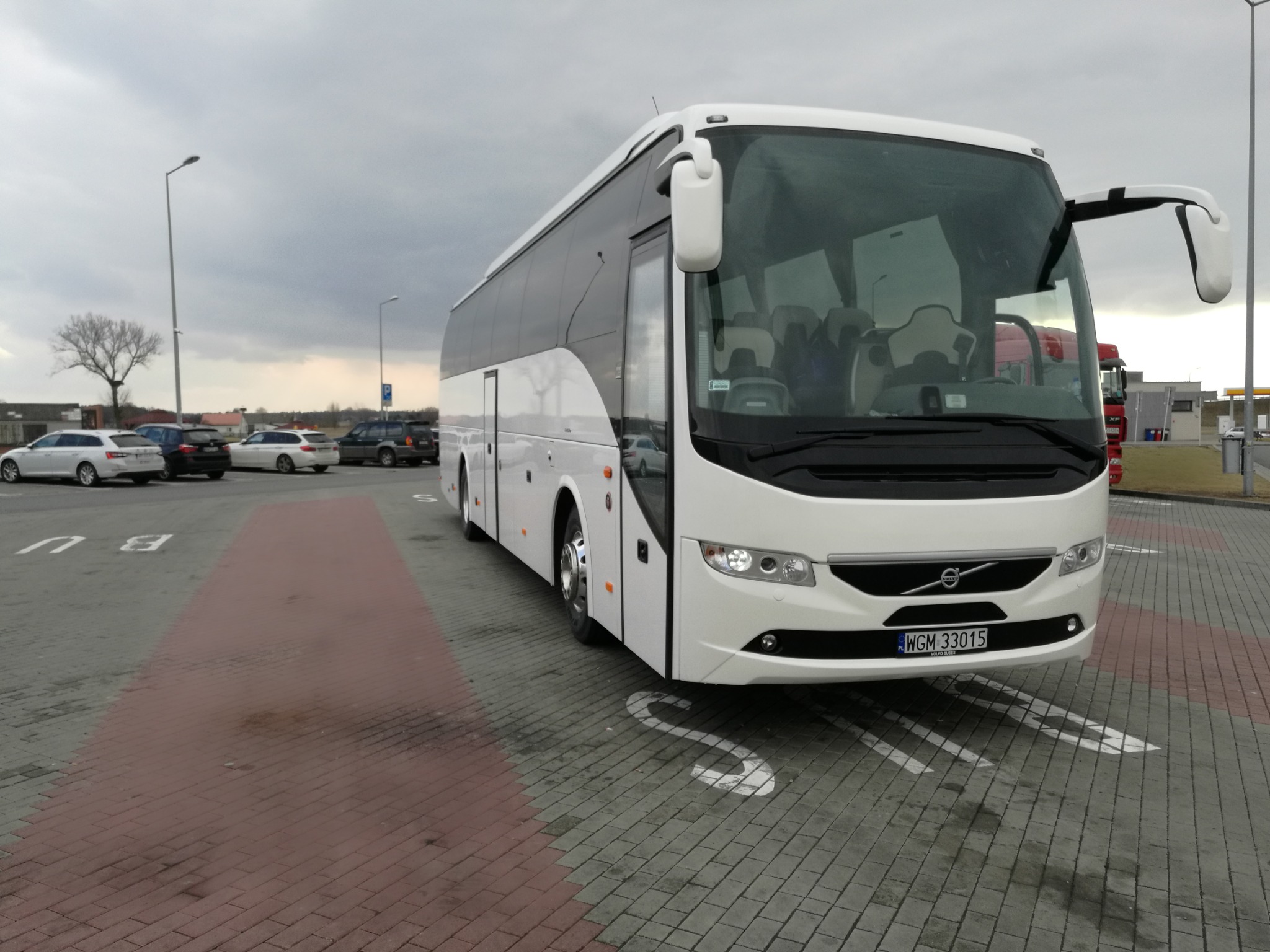 Biały autokar Volvo na parkingu w Warszawie, widoczny numer rejestracyjny WGM 33015, za nim zaparkowane samochody osobowe i ciężarówka.