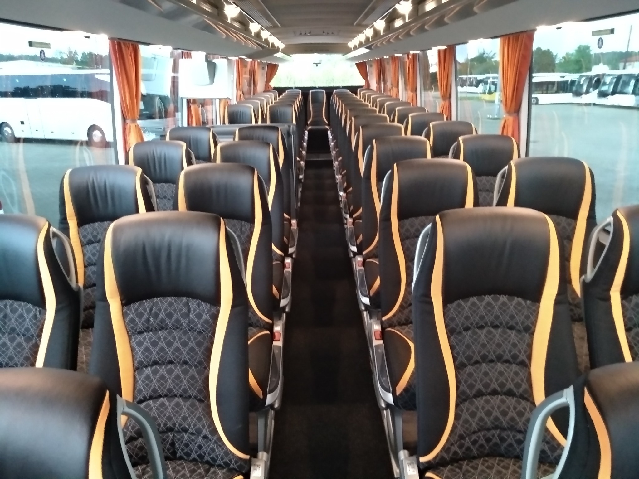 Wnętrze autobusu turystycznego z rzędami czarnych foteli z żółtymi wstawkami i wzorzystym obiciem, widok z przodu na puste przejście między fotelami, za oknami widoczne inne autobusy.