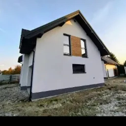 Nowoczesny dom jednorodzinny z białą elewacją i drewnianymi panelami dekoracyjnymi na piętrze, widoczny z perspektywy ogrodu w trakcie wykańczania.