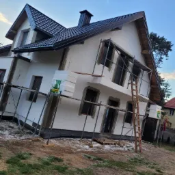 Dom jednorodzinny w trakcie prac związanych z dociepleniem elewacji, widoczne rusztowania, styropianowe płyty izolacyjne, okna zabezpieczone folią oraz drewniana drabina oparta o ścianę.