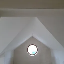 Wnętrze poddasza z oknem okrągłym w szczycie, ściany w trakcie wygładzania, sufit pomalowany na biało, widoczne ślady po pracach wykończeniowych.