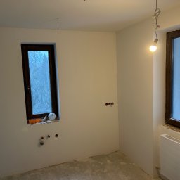 ŁUK-WIT Łukasz Witek - Pokój w trakcie remontu: świeżo pomalowane ściany na biało, dwa okna z brązowymi ramami, widoczne instalacje elektryczne i hydrauliczne, surowa podłoga, prowizoryczne oświetlenie żarówką.