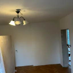 Wnętrze pokoju w trakcie odświeżania: świeżo pomalowane ściany na biało, parkiet w jodełkę, zabytkowa lampa sufitowa z białymi kloszami, drzwi w trakcie remontu, zabezpieczone folią meble.