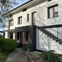 Dwukondygnacyjny dom z elewacją w odcieniach bieli i szarości, z zewnętrznymi schodami i ozdobną balustradą. Widoczne okna z brązowymi ramami i zadbana zieleń.