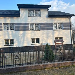 ŁUK-WIT Łukasz Witek - Dwupiętrowy dom z białymi ścianami i szarymi pasami, czarnym dachem i ozdobnym ogrodzeniem. Widoczne okna z odbiciami drzew i numer domu '42'.