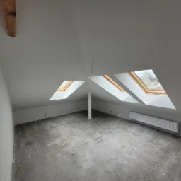 Poddasze po remoncie, z trzema oknami dachowymi, świeżo pomalowane ściany na biało, surowa betonowa podłoga, grzejnik zamontowany pod oknami.