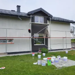 Dom jednorodzinny w trakcie malowania elewacji na biało i grafitowo, widoczne rusztowania, drabina, wiaderka z farbą i pracownik w kombinezonie malarskim.