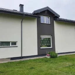 Dwukolorowa elewacja domu jednorodzinnego z szarą stolarką okienną i dachem, widoczne rynny, trawnik i fragment posesji.