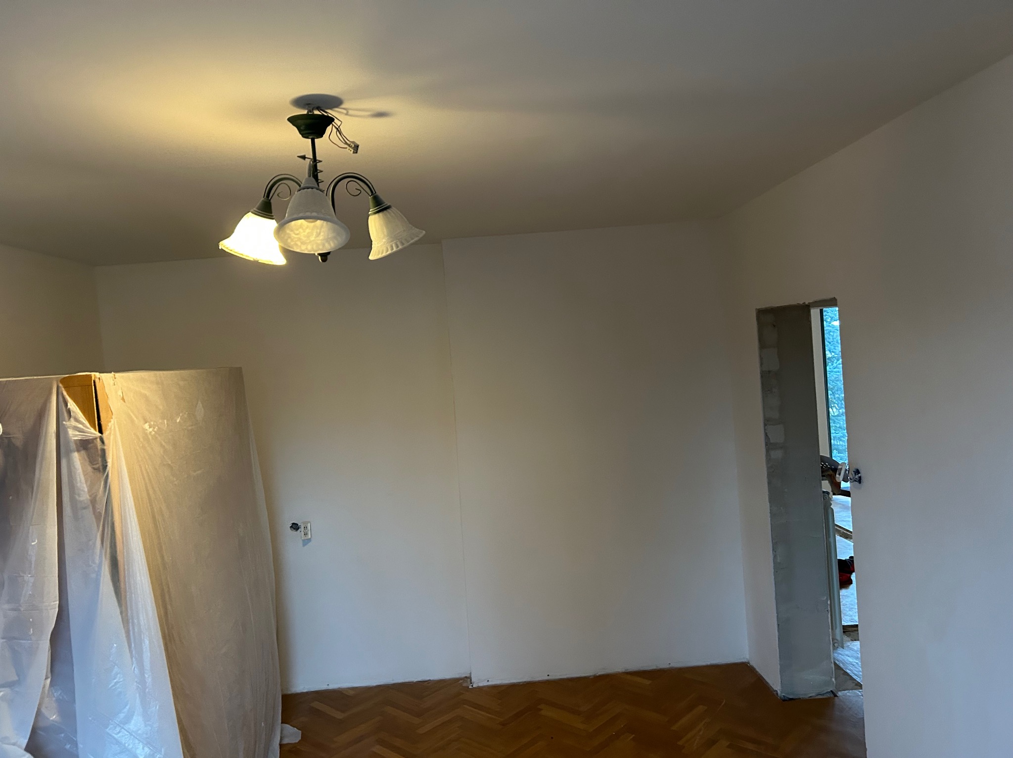 Wnętrze pokoju w trakcie odświeżania: świeżo pomalowane ściany na biało, parkiet w jodełkę, zabytkowa lampa sufitowa z białymi kloszami, drzwi w trakcie remontu, zabezpieczone folią meble.