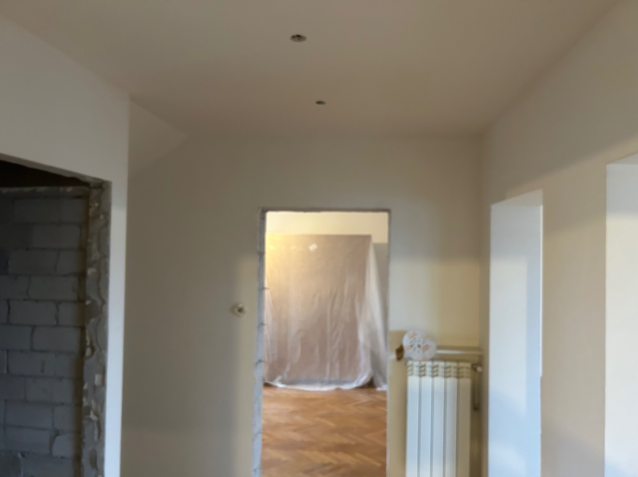 Wnętrze mieszkania w trakcie remontu: świeżo pomalowane ściany na biało, widoczne ślady po punktowym oświetleniu w suficie, fragment ściany z surowej cegły i zabezpieczone folią pomieszczenie...