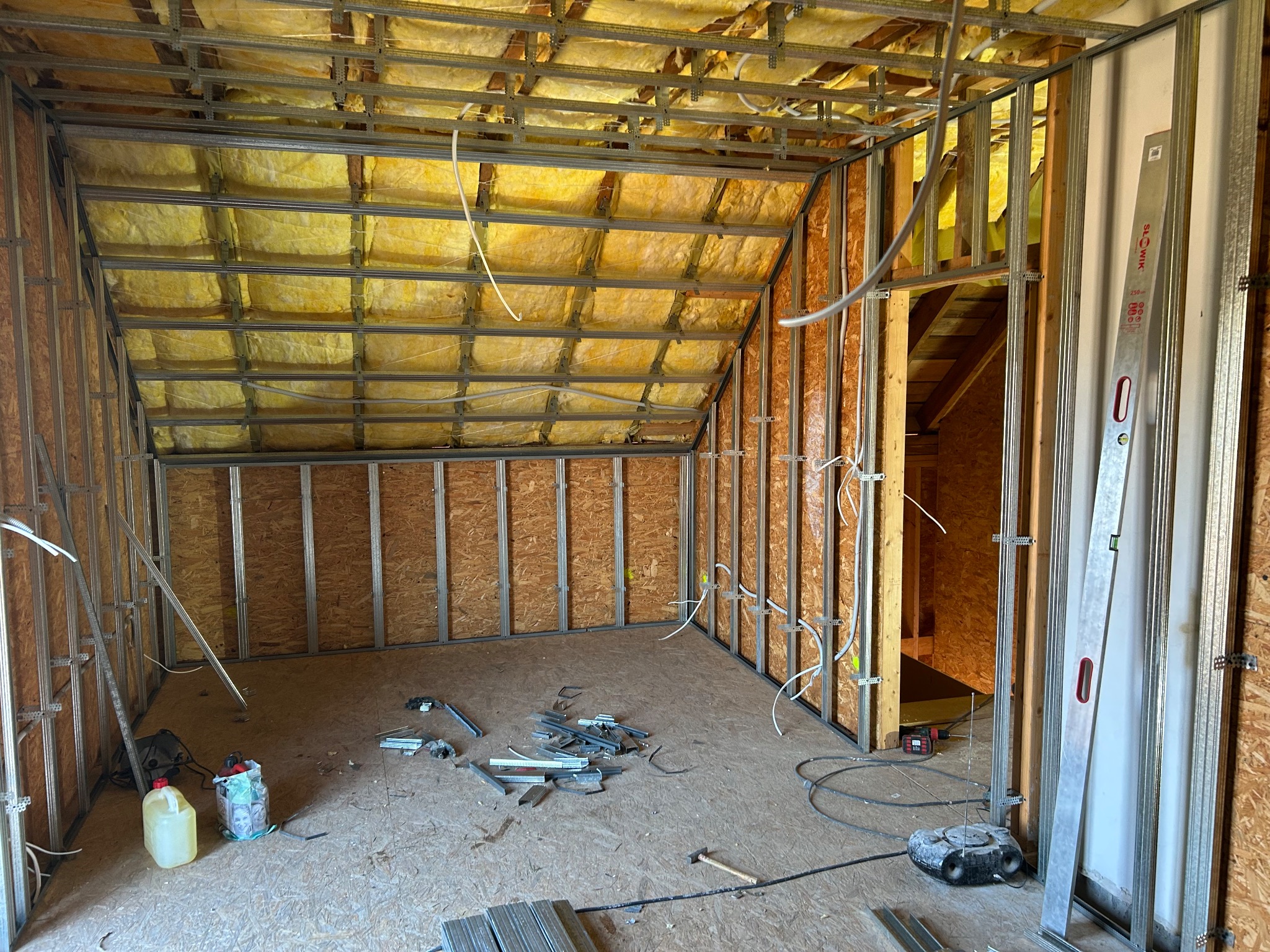Poddasze w trakcie adaptacji: widoczne metalowe profile, częściowo ułożona izolacja z wełny mineralnej, przewody elektryczne i narzędzia na podłodze. Etap prac wykończeniowych.