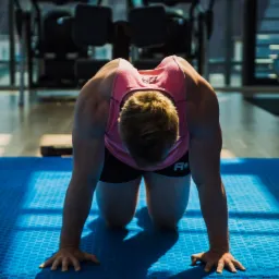 Mężczyzna w r&oacute;żowej koszulce i czarnych spodenkach ćwiczy na czworakach na niebieskiej macie w siłowni, z maszynami do ćwiczeń w tle.