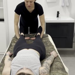 GROW UP STUDIO SPÓŁKA Z OGRANICZONĄ ODPOWIEDZIALNOŚCIĄ - Kobieta w czarnym stroju uciska stopy pacjentki leżącej na kozetce pokrytej brązowym materiałem imitującym skórę zwierzęcia, w tle umywalka z czarną szafką i podajnik na ręczniki.