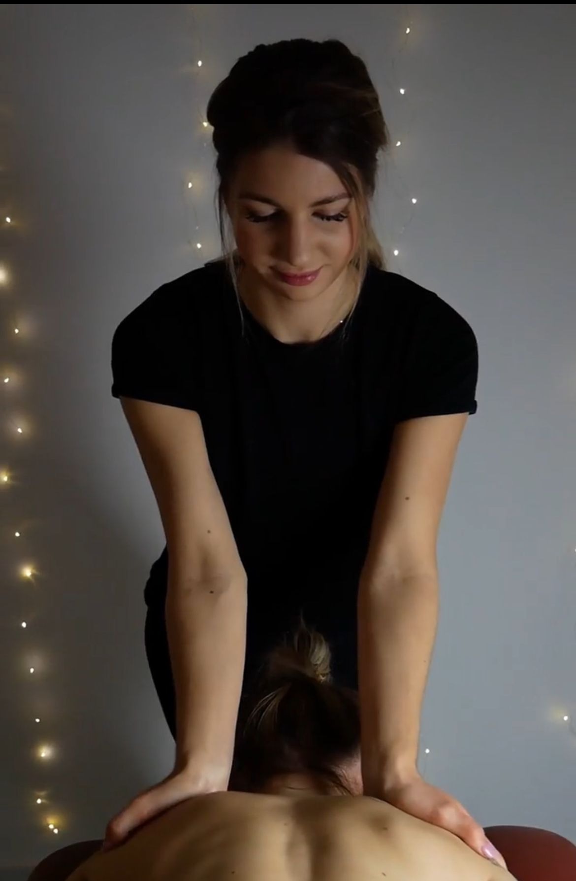 Kobieta w czarnej koszulce wykonuje masaż pleców innej kobiety na tle ściany z delikatnymi światełkami.