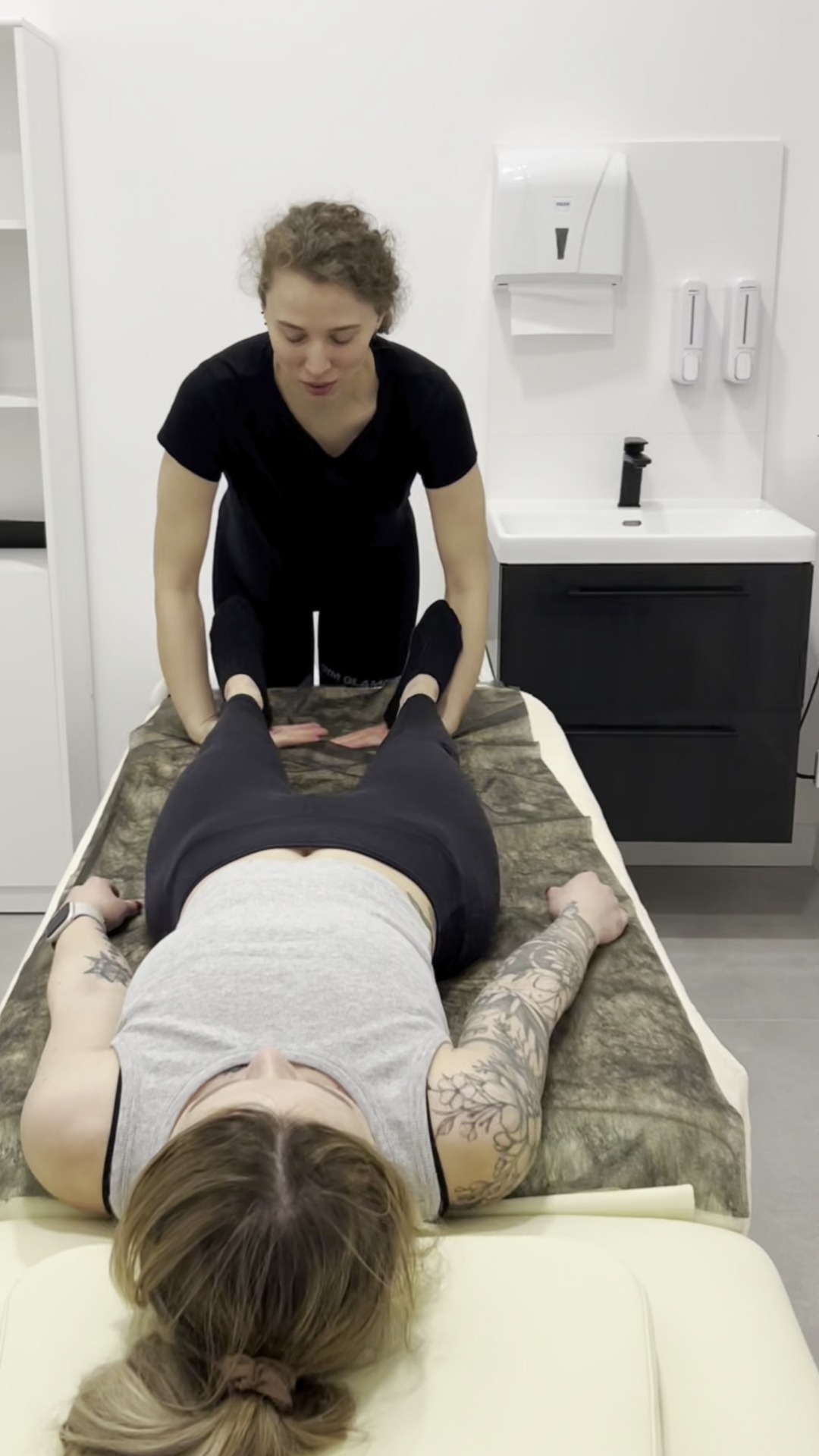 Kobieta w czarnym stroju uciska stopy pacjentki leżącej na kozetce pokrytej brązowym materiałem imitującym skórę zwierzęcia, w tle umywalka z czarną szafką i podajnik na ręczniki.