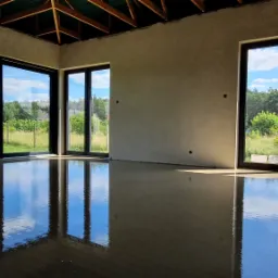 Świeżo wylana, gładka posadzka w jasnym pomieszczeniu z dużymi, czarnymi oknami, odbijającymi błękit nieba i zieleń drzew na zewnątrz.