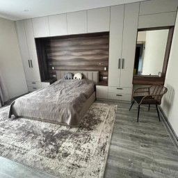 PRYIMENKO Sp. z o.o. - Sypialnia z zabudową na wymiar: jasne szafy zintegrowane z wezgłowiem łóżka z drewnianym panelem, dywan w odcieniach szarości, krzesło wiklinowe i toaletka z lustrem.