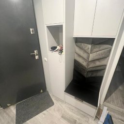 PRYIMENKO Sp. z o.o. - Wnęka z siedziskiem wykończona tapicerowanym panelem w kształcie chevronu, część zabudowy meblowej przedpokoju z szafkami i otwartą półką, widoczna czarna wycieraczka i drzwi wejściowe.