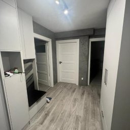 PRYIMENKO Sp. z o.o. - Przedpokój z szarą podłogą imitującą drewno, białą zabudową meblową z szafkami i siedziskiem, oraz dwiema białymi drzwiami na tle szarej ściany z dekoracyjnym wykończeniem.