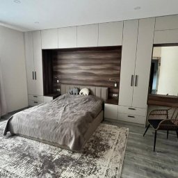 PRYIMENKO Sp. z o.o. - Szafa wnękowa z zabudową łóżka: jasne fronty, drewniana wnęka nad wezgłowiem, szara narzuta na łóżku, dywan w odcieniach szarości, krzesło z rattanu.