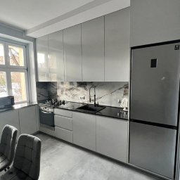 PRYIMENKO Sp. z o.o. - Nowoczesna kuchnia z szarymi, lakierowanymi frontami, czarnym blatem i marmurowym backsplashiem, oświetlona naturalnym światłem z okna.