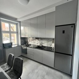 PRYIMENKO Sp. z o.o. - Nowoczesna kuchnia w odcieniach szarości z błyszczącymi frontami szafek, czarnym blatem i marmurowym backsplashiem. Widoczne krzesła przy stole, okno z widokiem na zewnątrz oraz wbudowana lodówka.