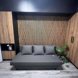 PRYIMENKO Sp. z o.o. - Szara sofa z trzema poduszkami na tle drewnianej ściany z pionowych listew i szafy z drewnianymi frontami, całość w nowoczesnym wnętrzu.