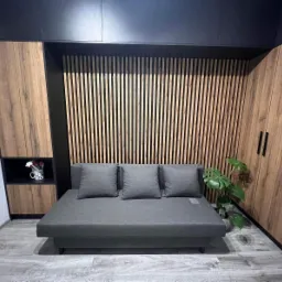 Szara sofa z trzema poduszkami na tle drewnianej ściany z pionowych listew i szafy z drewnianymi frontami, całość w nowoczesnym wnętrzu.